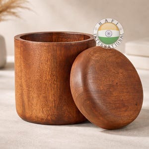 May include: A cylindrical wooden container with a matching lid. The container and lid are made of a warm-toned wood, likely mahogany or a similar species. The lid is oval-shaped and rests beside the container. The container is approximately 4 inches tall.