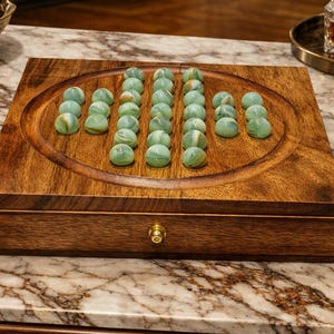 May include: A wooden solitaire game board with a drawer, featuring a carved oval playing area. The board is filled with 37 green and white marbles. The game is set on a marble countertop.
