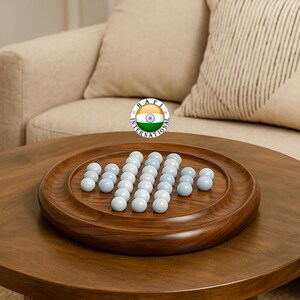 May include: A wooden solitaire game with light blue marbles arranged on a round, wooden tray. The game sits on a dark brown, round coffee table. The background includes a beige sofa and a decorative vase.