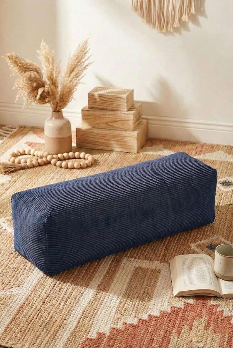 Może przedstawiać: Granatowa prostokątna poduszka sztruksowa, prawdopodobnie bolster do jogi, leży na wzorzystym dywanie. Poduszka jest w centrum, z wazonem z suszonymi roślinami i drewnianymi pudełkami w tle. W pobliżu książka i kubek.