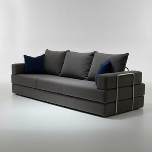 May include: A modern gray sofa with three gray cushions and two navy blue accent pillows. The sofa has a low profile and a metal frame detail on the armrest. The sofa is approximately 80 inches long.