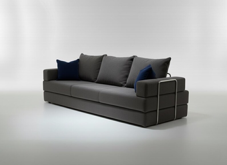May include: A modern gray sofa with three gray cushions and two navy blue accent pillows. The sofa has a low profile and a metal frame detail on the armrest. The sofa is approximately 80 inches long.