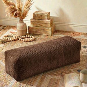 Może przedstawiać: Prostokątny, brązowy puf z sztruksu stoi na wzorzystym dywanie. Teksturowana powierzchnia pufa jest widoczna. W tle wazon z suszonymi roślinami, drewniane pudełka i książka z kubkiem tworzą przytulną atmosferę.