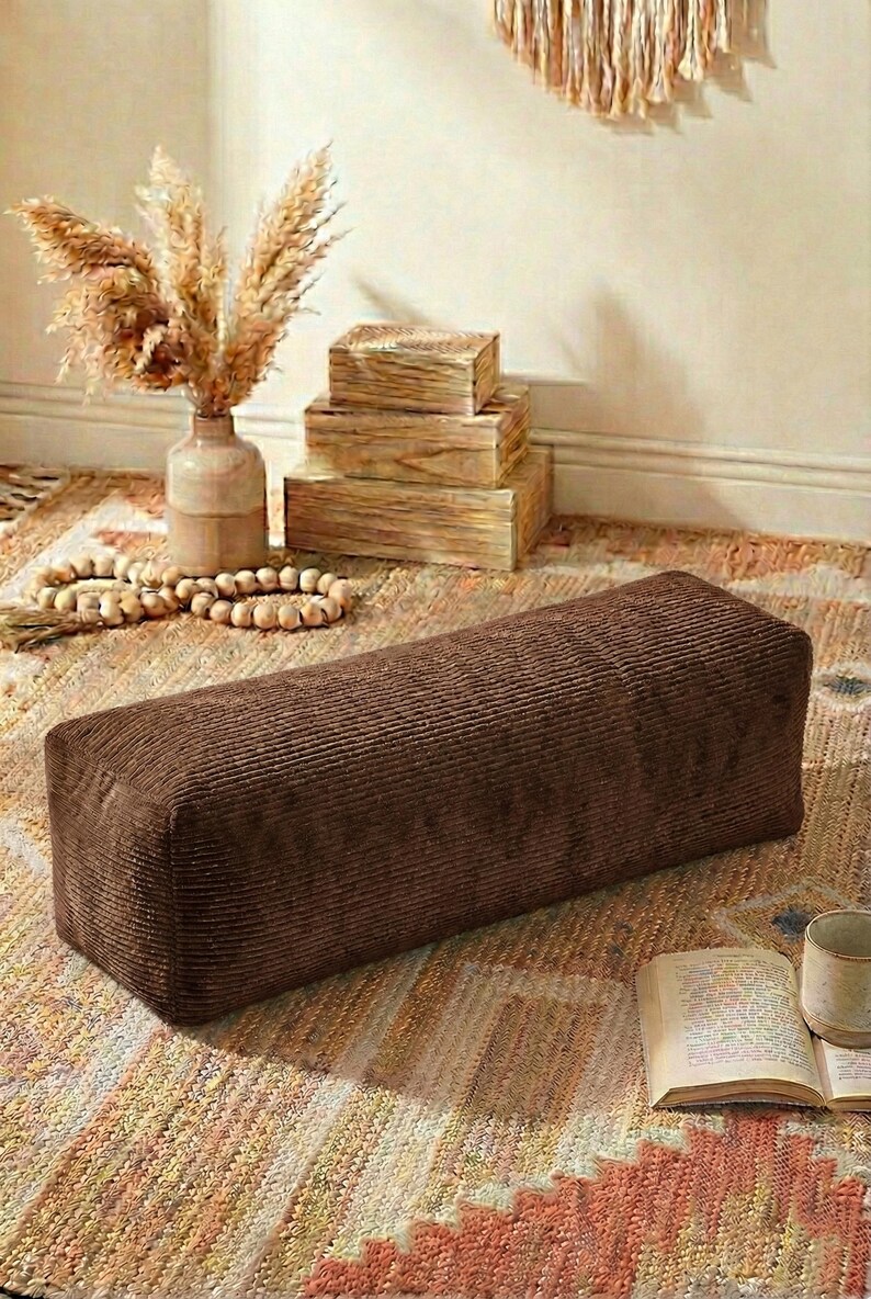 Może przedstawiać: Prostokątny, brązowy puf z sztruksu stoi na wzorzystym dywanie. Teksturowana powierzchnia pufa jest widoczna. W tle wazon z suszonymi roślinami, drewniane pudełka i książka z kubkiem tworzą przytulną atmosferę.