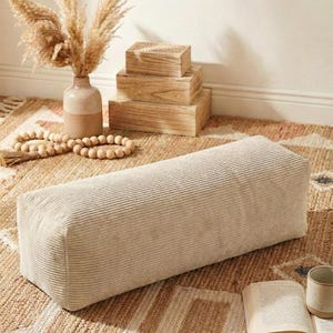 Może przedstawiać: Prostokątna, beżowa poduszka podłogowa z sztruksu. Poduszka leży na wzorzystym dywanie, z wazonem z suszonymi roślinami, drewnianymi pudełkami i girlandą z koralik&oacute;w w tle. Widoczna jest r&oacute;wnież książka i kubek.