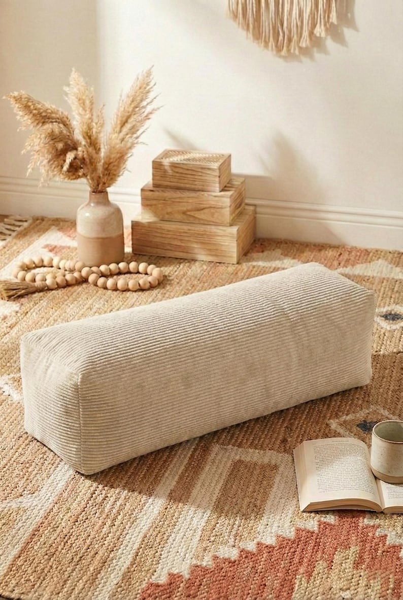 Może przedstawiać: Prostokątna, beżowa poduszka podłogowa z sztruksu. Poduszka leży na wzorzystym dywanie, z wazonem z suszonymi roślinami, drewnianymi pudełkami i girlandą z koralik&oacute;w w tle. Widoczna jest r&oacute;wnież książka i kubek.