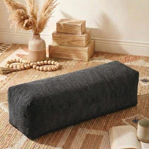 Może przedstawiać: Ciemnoszara, prostokątna poduszka podłogowa z sztruksu. Poduszka leży na wzorzystym dywanie o neutralnej palecie barw. Na zdjęciu znajduje się r&oacute;wnież wazon z suszonymi roślinami, drewniane pudełka i książka.