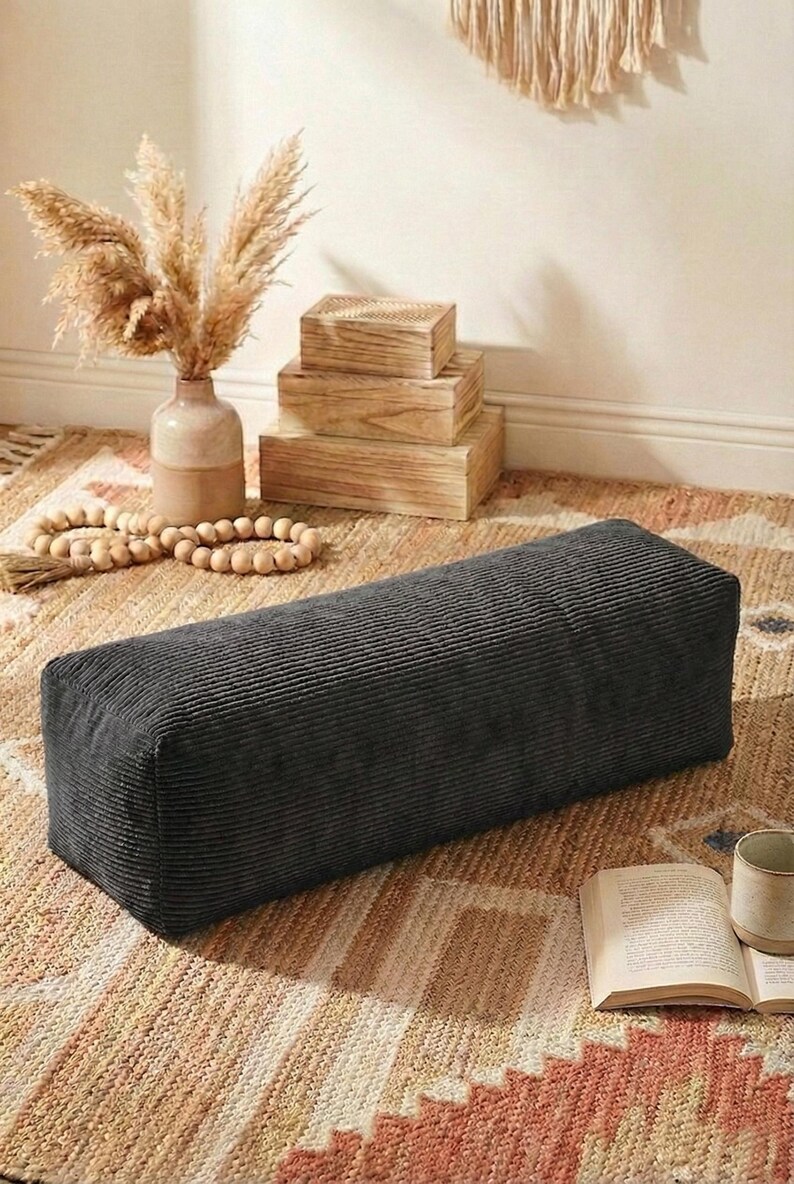 Może przedstawiać: Ciemnoszara, prostokątna poduszka podłogowa z sztruksu. Poduszka leży na wzorzystym dywanie o neutralnej palecie barw. Na zdjęciu znajduje się r&oacute;wnież wazon z suszonymi roślinami, drewniane pudełka i książka.