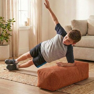 Może przedstawiać: Osoba wykonuje ćwiczenie planku bocznego na brązowej pufie sztruksowej. Osoba ma na sobie szarą i granatową koszulkę, czarne szorty i szare trampki. Ćwiczenie wykonywane jest na beżowej macie, z dywanem w neutralnych odcieniach i beżową sofą w tle.