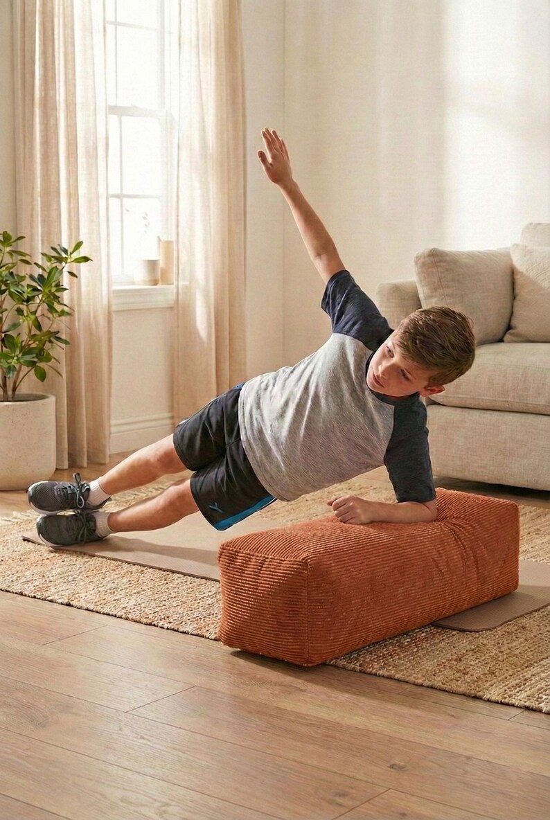 Może przedstawiać: Osoba wykonuje ćwiczenie planku bocznego na brązowej pufie sztruksowej. Osoba ma na sobie szarą i granatową koszulkę, czarne szorty i szare trampki. Ćwiczenie wykonywane jest na beżowej macie, z dywanem w neutralnych odcieniach i beżową sofą w tle.