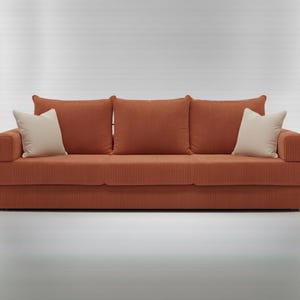 May include: A modern, orange corduroy sofa with three matching back cushions and two off-white accent pillows. The sofa has a low profile and a minimalist design, perfect for a living room or apartment.