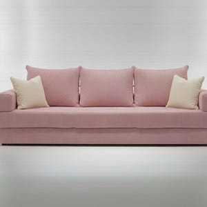 May include: A pink sofa with three matching back cushions and two cream-colored accent pillows. The sofa has a textured fabric and a low profile design, suitable for a living room or lounge area.