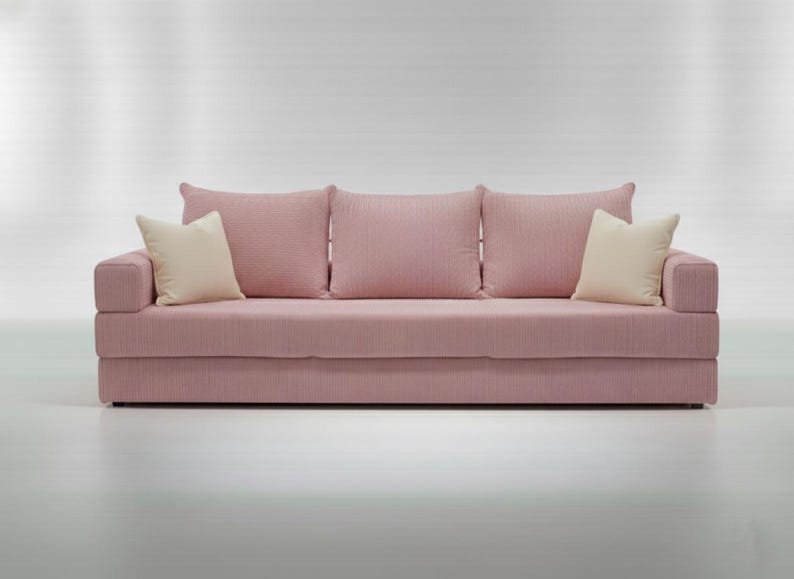May include: A pink sofa with three matching back cushions and two cream-colored accent pillows. The sofa has a textured fabric and a low profile design, suitable for a living room or lounge area.