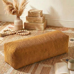 Może przedstawiać: Prostokątny puf z sztruksu w kolorze musztardowym. Puf stoi na wzorzystym dywanie, z wazonem z suszonymi kwiatami, drewnianymi pudełkami i książką. Tematem przewodnim jest wystr&oacute;j domu.