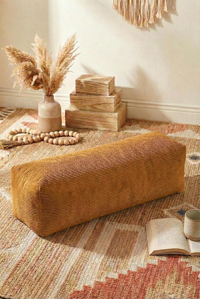 Może przedstawiać: Prostokątny puf z sztruksu w kolorze musztardowym. Puf stoi na wzorzystym dywanie, z wazonem z suszonymi kwiatami, drewnianymi pudełkami i książką. Tematem przewodnim jest wystr&oacute;j domu.