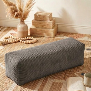 Może przedstawiać: Prostokątna, szara pufa sztruksowa stoi na wzorzystym dywanie. Na zdjęciu znajduje się r&oacute;wnież wazon z suszonymi roślinami, drewniane pudełka oraz książka z kubkiem. Scena przywodzi na myśl przytulną przestrzeń mieszkalną w neutralnych odcieniach.