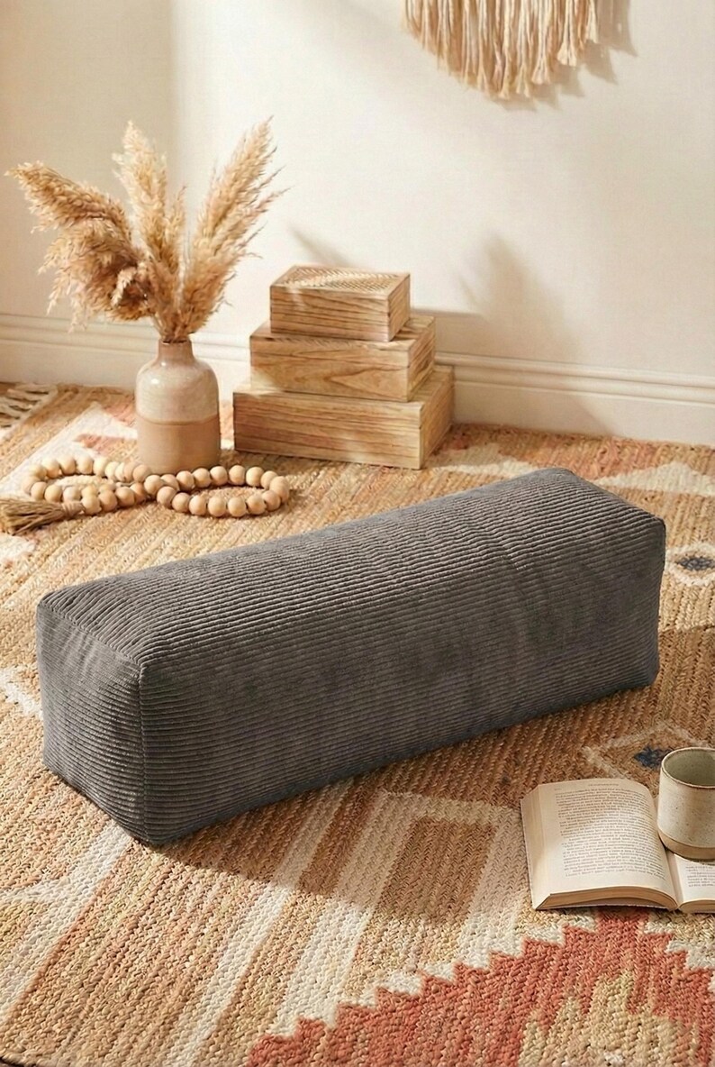 Może przedstawiać: Prostokątna, szara pufa sztruksowa stoi na wzorzystym dywanie. Na zdjęciu znajduje się r&oacute;wnież wazon z suszonymi roślinami, drewniane pudełka oraz książka z kubkiem. Scena przywodzi na myśl przytulną przestrzeń mieszkalną w neutralnych odcieniach.