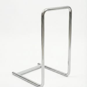 May include: A minimalist, chrome-finished metal stand. The stand features a rectangular frame supported by a horizontal base. The design is simple and modern, suitable for displaying items.