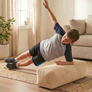 Może przedstawiać: Osoba wykonuje ćwiczenie planku bocznego na beżowym bloku do ćwiczeń sztruksowych. Osoba ma na sobie szarą i granatową koszulkę, czarne szorty i szare trampki. Ćwiczenie wykonywane jest na brązowej macie na tkanej macie.