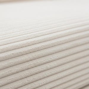 May include: Close-up of a beige corduroy fabric. The fabric has a ribbed texture with parallel lines running across the surface. The color is a neutral, light beige.