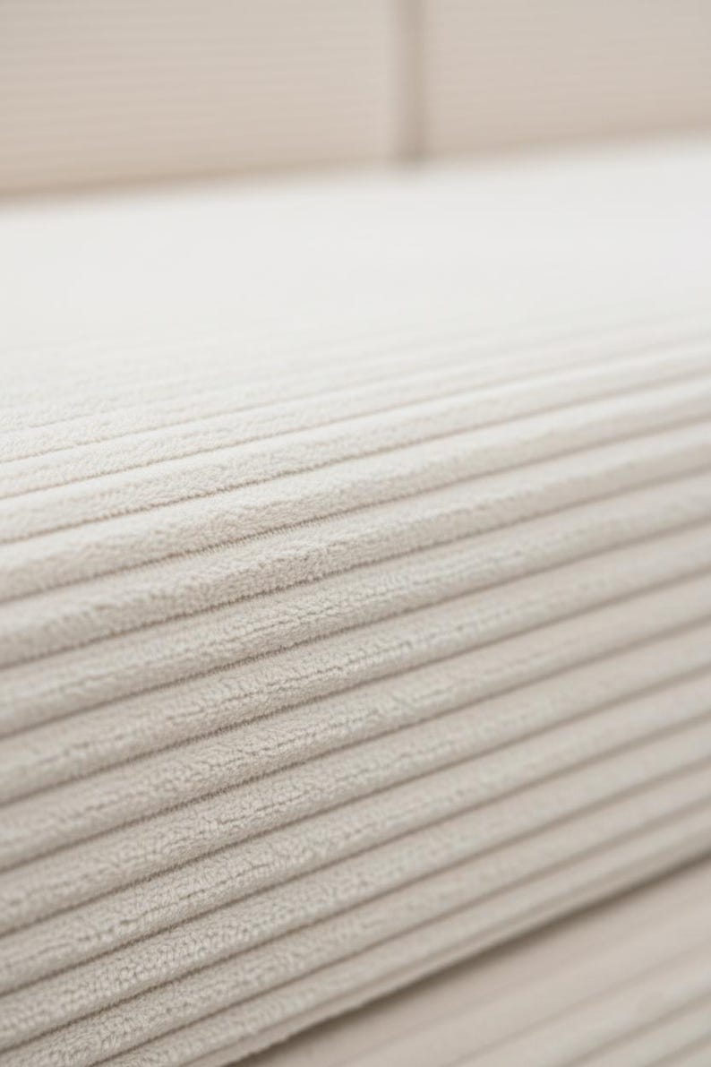 May include: Close-up of a beige corduroy fabric. The fabric has a ribbed texture with parallel lines running across the surface. The color is a neutral, light beige.