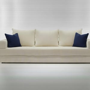 May include: A cream-colored sofa with three matching back cushions and two navy blue throw pillows. The sofa has a simple, modern design and is set against a white background. The sofa's dimensions are approximately 80 inches wide.