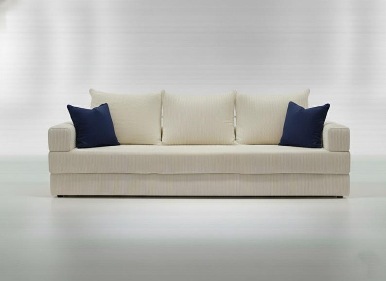May include: A cream-colored sofa with three matching back cushions and two navy blue throw pillows. The sofa has a simple, modern design and is set against a white background. The sofa's dimensions are approximately 80 inches wide.