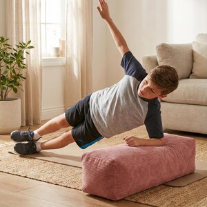 Może przedstawiać: Osoba wykonuje ćwiczenie plank boczny na r&oacute;żowej poduszce z sztruksu. Osoba ma na sobie szarą i granatową koszulkę z kr&oacute;tkim rękawem, czarne szorty i szare trampki. Poduszka leży na brązowej macie, a w tle widać okno i beżową sofę.