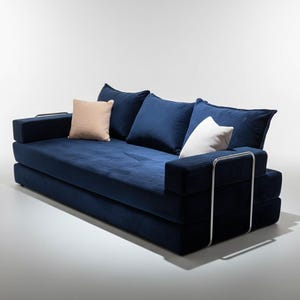May include: A modern, navy blue sofa with three large, matching pillows and two smaller accent pillows. The sofa has a low profile and a minimalist design, with chrome arm supports.