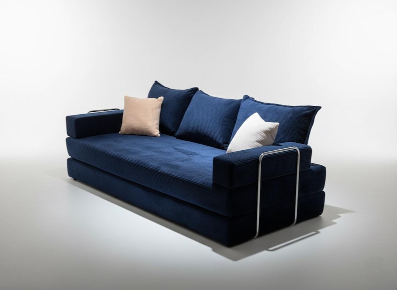 May include: A modern, navy blue sofa with three large, matching pillows and two smaller accent pillows. The sofa has a low profile and a minimalist design, with chrome arm supports.
