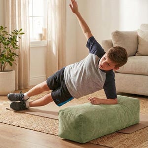 Może przedstawiać: Osoba wykonuje ćwiczenie w pozycji bocznej deski, opierając się na zielonym bloku sztruksowym. Ma na sobie szaro-granatową koszulkę i czarne szorty. Ćwiczenie wykonywane jest na brązowej macie do jogi, z neutralnym tłem.