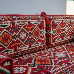 Puede incluir: Un sofá rojo estampado con cojines a juego. Los cojines y el sofá presentan un diseño geométrico en blanco, verde, naranja y negro. El sofá tiene un borde rojo y borlas.