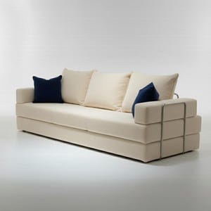 May include: A modern, cream-colored sofa with three off-white cushions and two navy blue accent pillows. The sofa features a geometric armrest design with metal accents, creating a minimalist aesthetic.