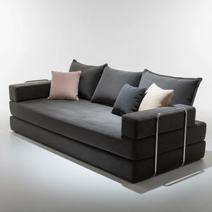 May include: A modern, dark gray sofa with multiple cushions in varying shades of gray and a blush pink accent pillow. The sofa has a low profile and a minimalist design, with chrome accents on the arms.