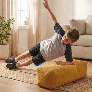 Może przedstawiać: Osoba wykonująca ćwiczenie planku bocznego na musztardowej ż&oacute;łtej ławce sztruksowej. Osoba ma na sobie szarą i granatową koszulkę z kr&oacute;tkim rękawem i czarne szorty. Ćwiczenie wykonywane jest na brązowej macie do jogi, z neutralnym dywanem i beżową sofą w tle.