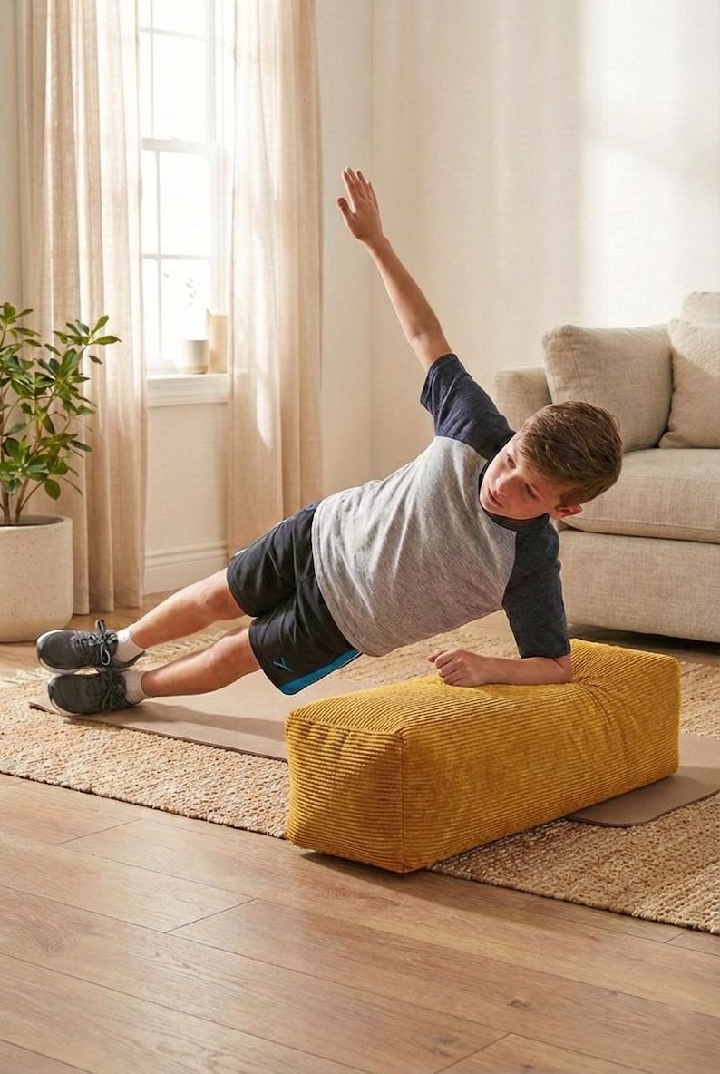 Może przedstawiać: Osoba wykonująca ćwiczenie planku bocznego na musztardowej ż&oacute;łtej ławce sztruksowej. Osoba ma na sobie szarą i granatową koszulkę z kr&oacute;tkim rękawem i czarne szorty. Ćwiczenie wykonywane jest na brązowej macie do jogi, z neutralnym dywanem i beżową sofą w tle.