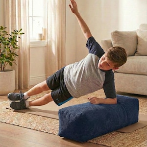 Może przedstawiać: Osoba wykonuje ćwiczenie planku bocznego na granatowym, prostokątnym bloku do ćwiczeń o fakturze sztruksu. Osoba ma na sobie szarą i granatową koszulkę z kr&oacute;tkim rękawem oraz czarne szorty. Ćwiczenie wykonywane jest na brązowej macie, z dywanem i sofą w neutralnych odcieniach w tle.