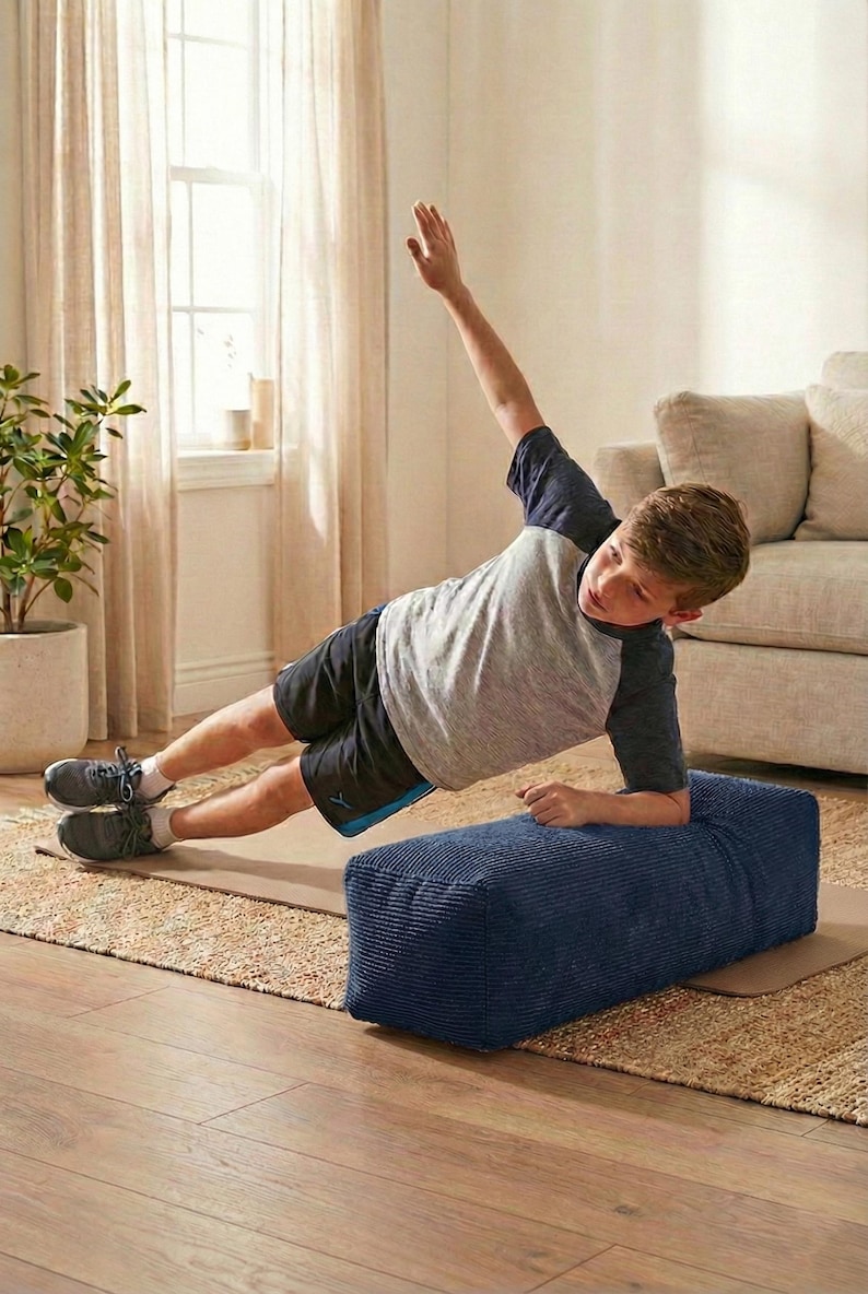 Może przedstawiać: Osoba wykonuje ćwiczenie planku bocznego na granatowym, prostokątnym bloku do ćwiczeń o fakturze sztruksu. Osoba ma na sobie szarą i granatową koszulkę z kr&oacute;tkim rękawem oraz czarne szorty. Ćwiczenie wykonywane jest na brązowej macie, z dywanem i sofą w neutralnych odcieniach w tle.