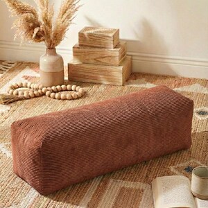 Może przedstawiać: Prostokątna pufa z sztruksu w ciepłym kolorze terakoty. Pufa stoi na wzorzystym dywanie, obok wazonu z suszoną trawą pampasową, drewnianych pudełek i książki. Całość tworzy przytulną i zachęcającą atmosferę.