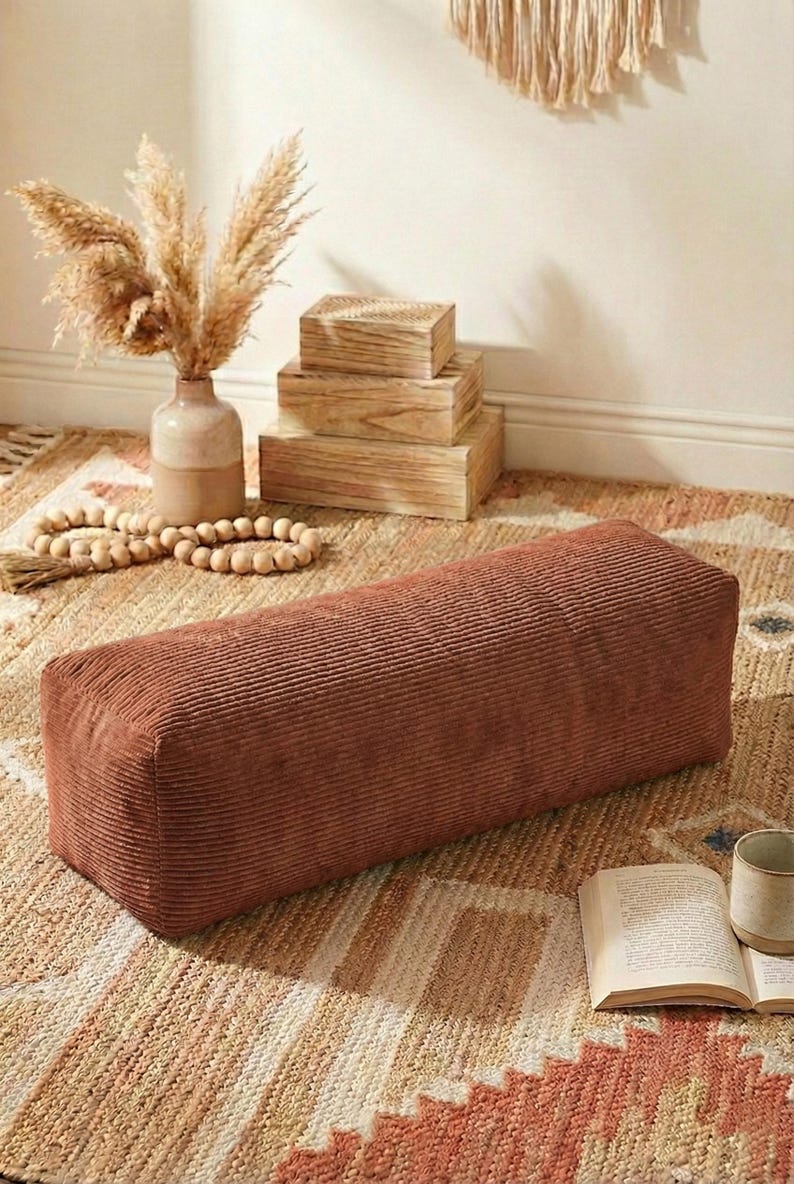 Może przedstawiać: Prostokątna pufa z sztruksu w ciepłym kolorze terakoty. Pufa stoi na wzorzystym dywanie, obok wazonu z suszoną trawą pampasową, drewnianych pudełek i książki. Całość tworzy przytulną i zachęcającą atmosferę.