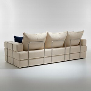 May include: A modern, cream-colored sofa with a geometric design. The sofa features a series of stacked rectangular blocks and a metal frame. It has three large, soft back cushions and a single navy blue accent pillow.