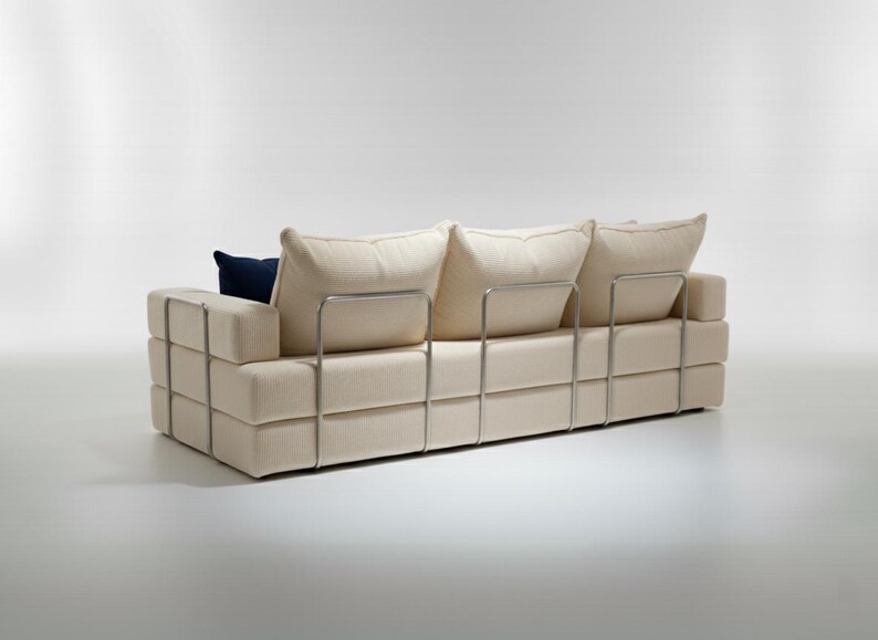 May include: A modern, cream-colored sofa with a geometric design. The sofa features a series of stacked rectangular blocks and a metal frame. It has three large, soft back cushions and a single navy blue accent pillow.