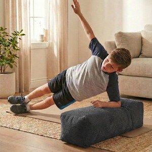 Może przedstawiać: Osoba wykonuje ćwiczenie w postaci deski bocznej, opierając się na szarym, teksturowanym bloku. Ma na sobie szaro-granatową koszulkę, czarne szorty i szare trampki. Ćwiczenie odbywa się na macie na dywanie.