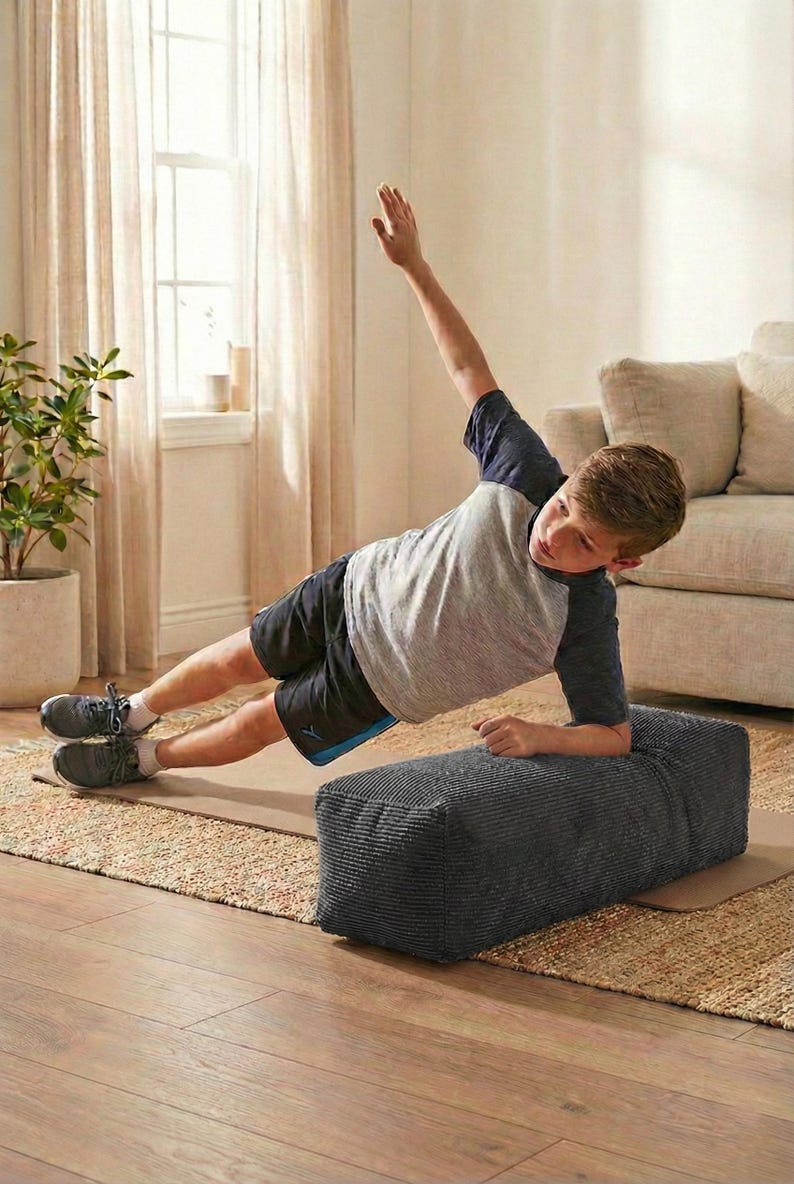 Może przedstawiać: Osoba wykonuje ćwiczenie w postaci deski bocznej, opierając się na szarym, teksturowanym bloku. Ma na sobie szaro-granatową koszulkę, czarne szorty i szare trampki. Ćwiczenie odbywa się na macie na dywanie.