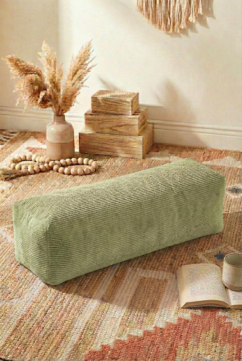 Może przedstawiać: Prostokątny puf z sztruksu w kolorze szałwii. Puf stoi na wzorzystym dywanie, z wazonem z suszonymi kwiatami, drewnianymi pudełkami i książką w pobliżu. Paleta kolor&oacute;w jest neutralna i ziemista.