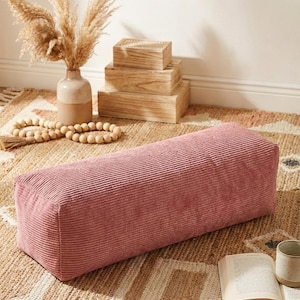 Może przedstawiać: Prostokątna, zakurzona r&oacute;żowa poduszka z sztruksu spoczywa na wzorzystym dywanie. Poduszka jest punktem centralnym, z wazonem z suszonymi kwiatami i drewnianymi pudełkami w tle. W pobliżu znajduje się książka i kubek.