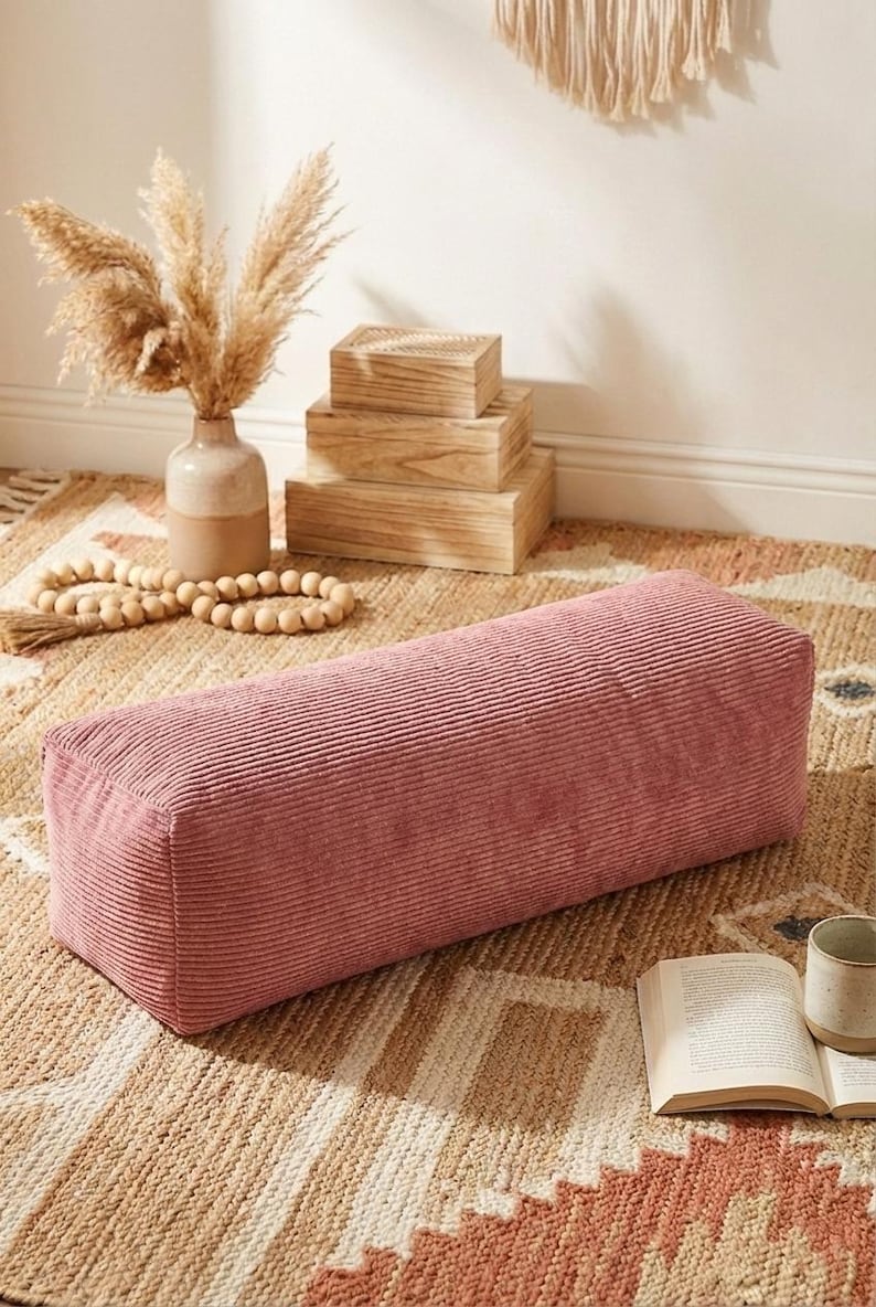 Może przedstawiać: Prostokątna, zakurzona r&oacute;żowa poduszka z sztruksu spoczywa na wzorzystym dywanie. Poduszka jest punktem centralnym, z wazonem z suszonymi kwiatami i drewnianymi pudełkami w tle. W pobliżu znajduje się książka i kubek.