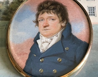 Op de afbeelding: Een miniatuurportret van een man in een blauwe jas met gouden knopen. Het portret is ingelijst in een gouden ovale lijst met een lus aan de bovenkant om op te hangen. De handtekening van de kunstenaar, "Chalon", is zichtbaar in de linkerbenedenhoek.