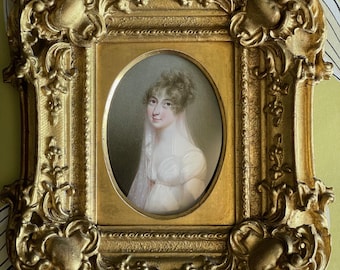 Puede incluir: Un retrato pintado de una persona con cabello oscuro, vistiendo un vestido blanco y un velo transparente, colocado en un marco ovalado dentro de un marco rectangular dorado, ornamentado con intrincados detalles de volutas y relieves.