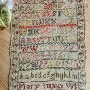 Puede incluir: Antiguo sampler con bordado de punto de cruz colorido. El textil presenta filas de letras, números y el nombre "Mary Imms" y el año "1812". El sampler tiene un borde decorativo sobre un fondo de lino beige.